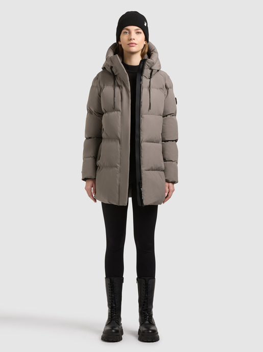Model trägt eine taupefarbene Steppjacke mit Kapuze, schwarzer Mütze und schwarzen Stiefeln.