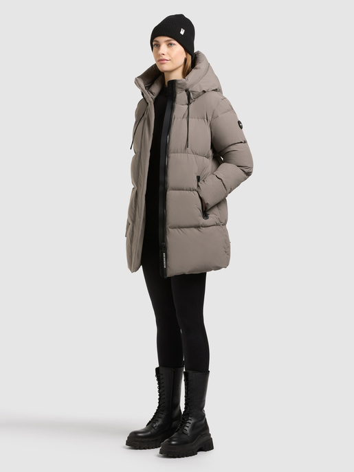 Model trägt eine taupefarbene Steppjacke mit Kapuze, schwarze Mütze und Schnürstiefel.