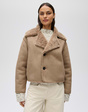 Model trägt eine beigefarbene kurze Jacke in Velourslederoptik mit breitem Teddyfutter-Kragen und dunklen Knöpfen.